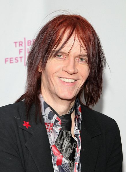 Nick Zedd smiling, with long hair, wearing a black coat with a red star on it over a multi-colored long sleeve, and a black and white necktie.