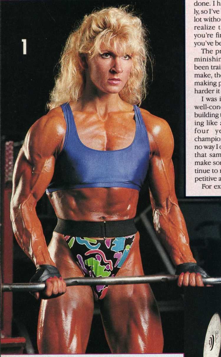 Nikki Fuller carrying a barbell while wearing a blue sports bra and a colorful panty