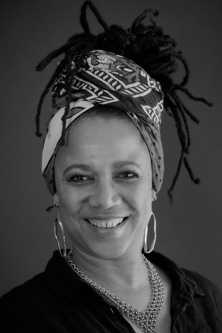 Nimmy March smiles while wearing a black collared shirt, pair of earrings, and necklaces