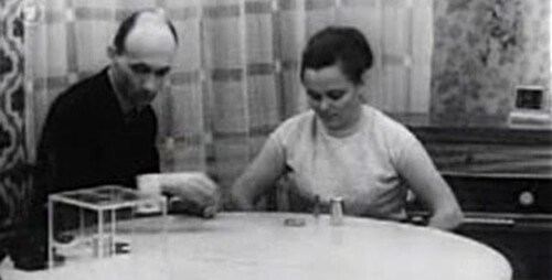 Nina Kulagina sitting and facing the objects placed on a round table, with curtains on the background, a clip from 1967 Telekinesis Experiment Russia, she is wearing a dress beside her is Eduard Naumo holding an object and placing it on the table wearing a white long sleeve under a black coat