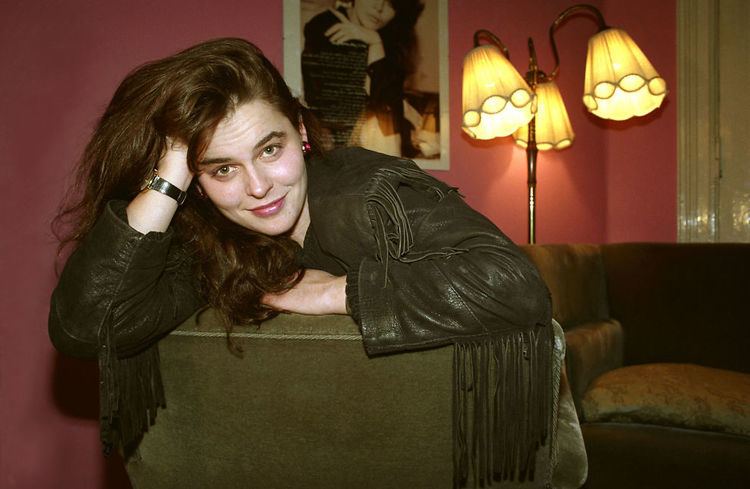 Nini Stoltenberg smiling while leaning on the couch having her hand in her hair with a lampstand and poster in the background and wearing a black coat and a black watch