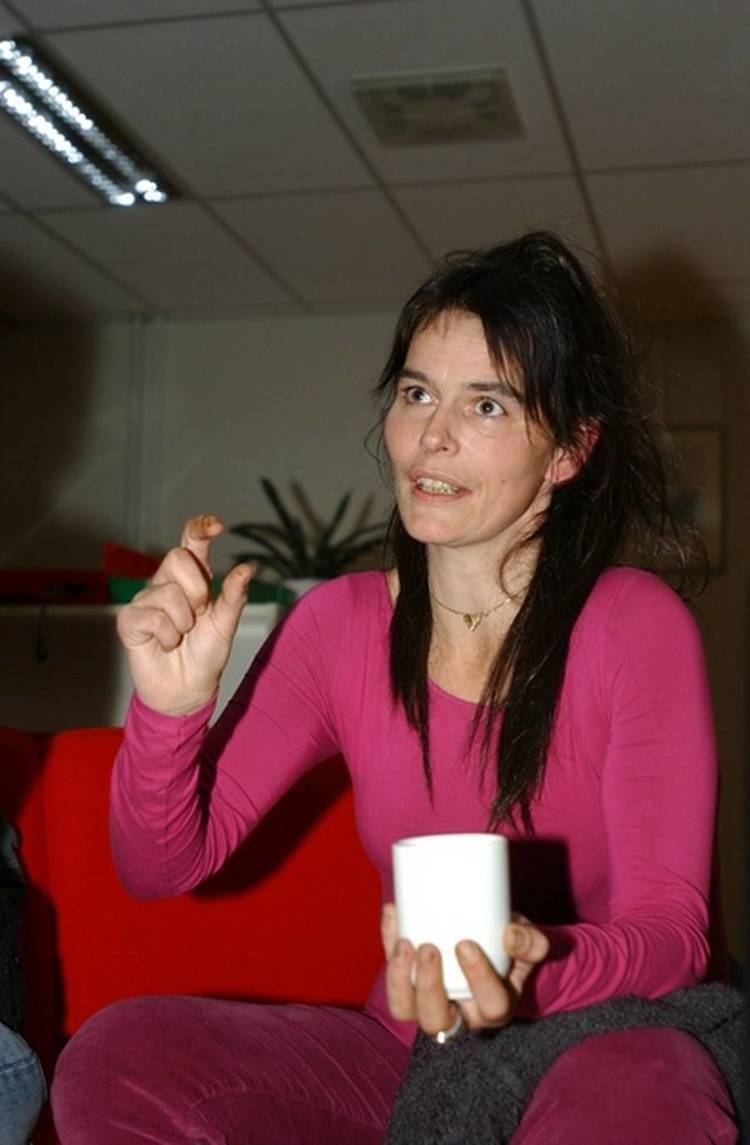 Nini Stoltenberg talking with someone having a mucky finger and holding a cup with her right hand while wearing a pink blouse and pants, a silver ring, a golden heart necklace and a ring