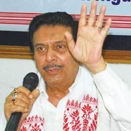 In a room with a large frame at the wall, Nipon Goswami is serious, sitting speaking while holding the mic with his right hand, and left hand up open, has black hair, mustache wearing silver rings and gold with red gem ring and a white long sleeve with red broided pattern.