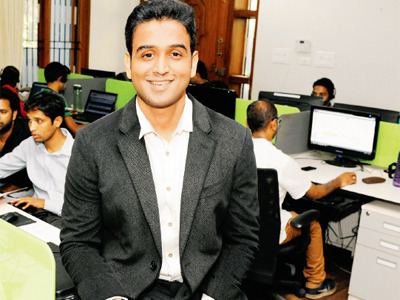 Nithin Kamath with a smiling face, wearing a black suit, and a white shirt with people at his back on their computers.