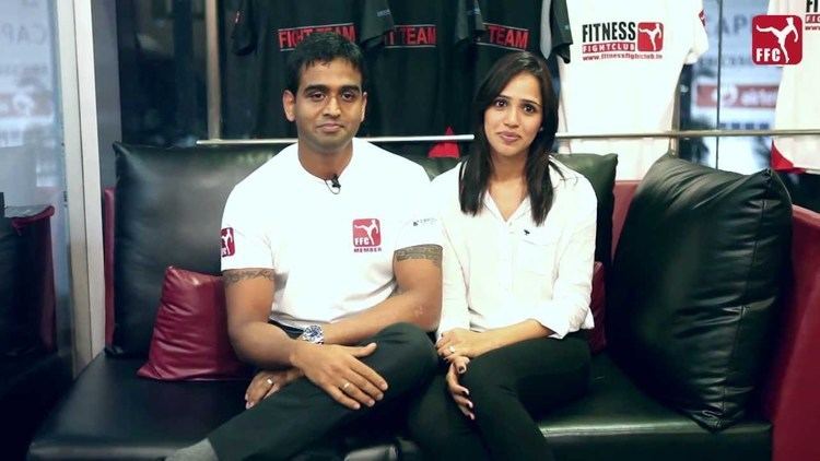 Nithin Kamath wearing a white polo shirt, a watch, rings, and black pants with his wife, Seema Patil wearing white long sleeves, a ring, and black pants, both are sitting on a black couch with shirts on their back.