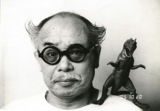 Nobuyoshi Araki with a mustache, wearing eyeglasses and a white shirt with a dinosaur toy on his shoulder.