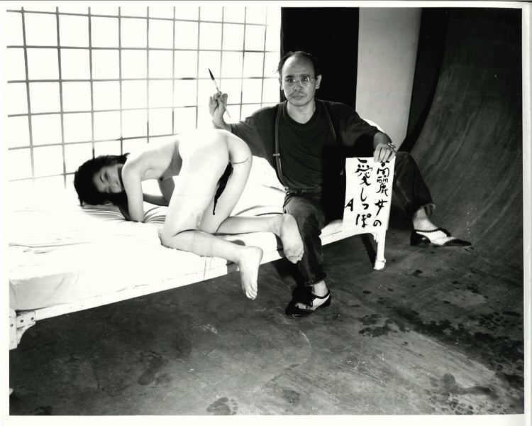 Nobuyoshi Araki wearing eyeglasses, a black shirt, and black jeans with a naked woman lying in the bed.