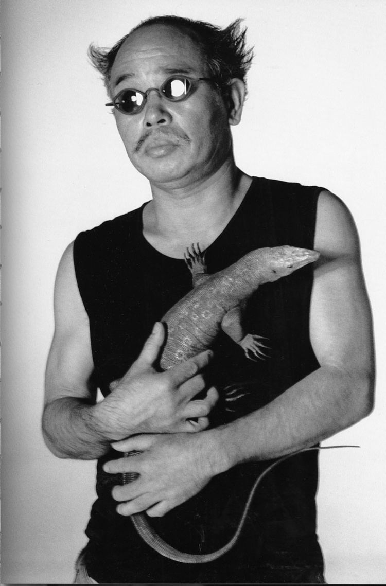 Nobuyoshi Araki wearing sunglasses and a black shirt while holding a big lizard.