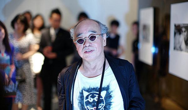 Nobuyoshi Araki wearing sunglasses, a black suit, and a white shirt with a cat printed on it.