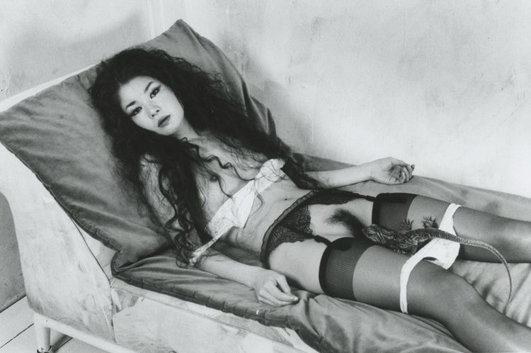 A woman lying in bed, with long curly hair, showing her boobs and wearing lingerie with a lizard on her top.