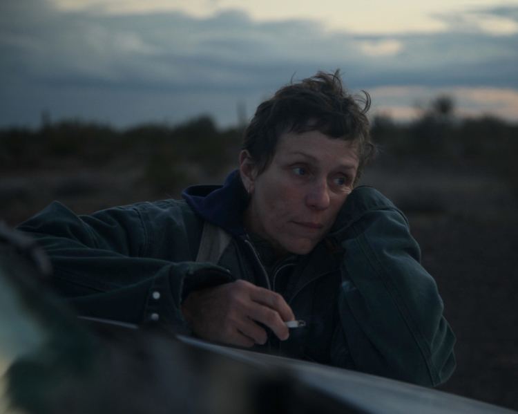 Frances McDormand holding a cigarette while looking at something and wearing a blue jacket in a scene from the 2020 American drama film, Nomadland