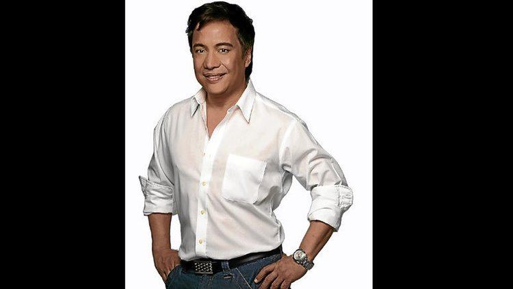 Nonoy Zuniga smiling and wearing a white buttoned polo shirt and jean pants with a black belt and a watch on his wrist.