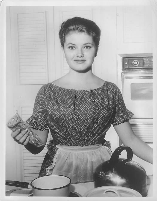 Noreen Corcoran is smiling, has black hair, and standing inside the kitchen, her right hand holding a pot holder, in front is a white bowl, a kettle, and a casserole, behind her(left) is a stove oven, she is wearing a black dress with buttons, white designs, and a white apron.