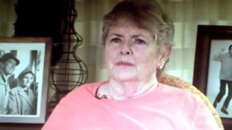 Noreen Corcoran is serious and has white hair, and behind her (on right) is a picture frame and a brown holder, and on left is also a picture frame, she is sitting down on a red chair, wearing earrings and a pink shirt.