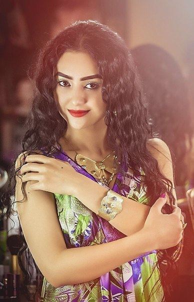 Noziya Karomatullo with a tight-lipped smile and long curly hair while wearing a green and violet sleeveless blouse, necklace, and bracelet