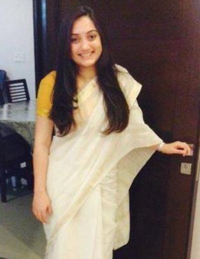 Nupur Sharma is smiling and holding the doorknob in her left hand with a dining table in the background, has black hair, a bindi on her forehead, wearing a bracelet and a yellow and white saree