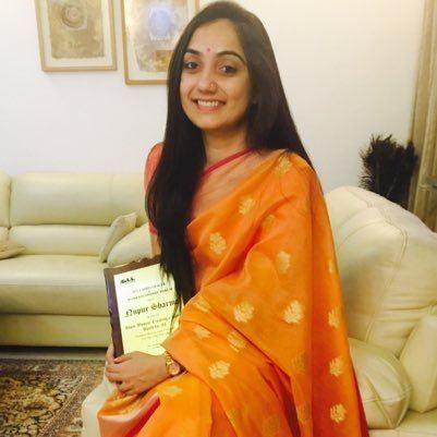 Nupur Sharma is smiling while sitting on a white couch and holding a certificate, with 2 frames on the wall in the background, she has black hair, a bindi on her forehead, a nose piercing, a watch on her left wrist, and an orange saree