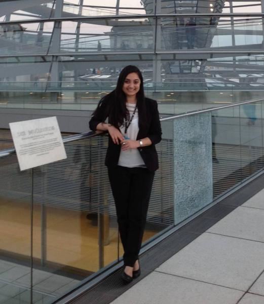 Nupur Sharma is smiling while leaning on her right arm in a glass hallway, she has black hair, wearing an ID strap on her neck, bracelet on her left hand and a ring on her left finger, black shoes, a white shirt under a black coat, and a black pants