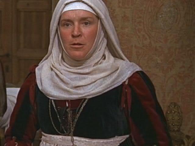Pat Heywood as Juliet's nurse looking serious in a scene from the 1968 film "Romeo and Juliet", wearing a black and red dress, white apron, necklace, and a white headdress