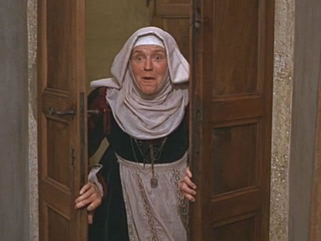 Pat Heywood as Juliet's nurse looking surprised while opening a door in a scene from the 1968 film "Romeo and Juliet", wearing a black and red dress, white apron, necklace and a white headdress