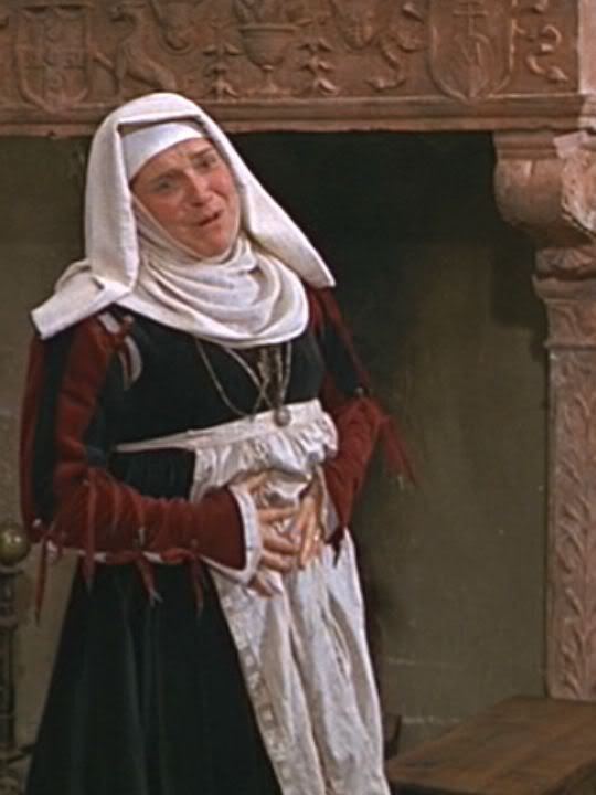 Pat Heywood as Juliet's nurse smiling in a scene from the 1968 film "Romeo and Juliet", wearing a black and red dress, white apron, necklace, and a white headdress