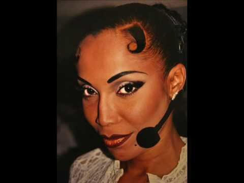 Octavia St. Laurent is serious, has black hair with side bangs on left, a mole on the lower left cheek, and a mic lapel, wearing silver earrings and a white top.