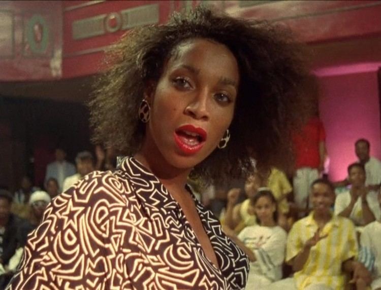 Octavia St Laurent is serious, has black curly hair, a mole on her right cheek, mouth half opened, behind her are different guests, she is wearing silver earrings and a white-brown top cleavage showing stars and crescent design.