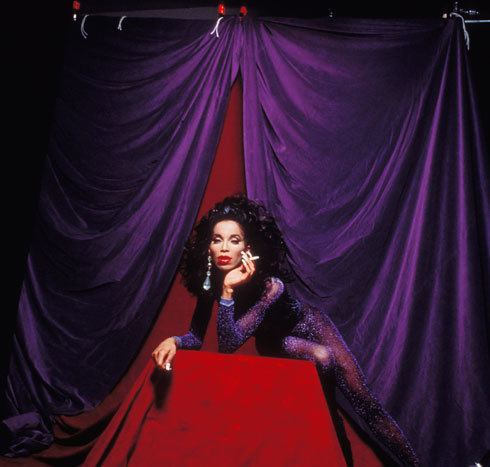 Octavia St Laurent is serious, has black curly hair, sitting down on a red cube platform, behind is a violet curtain, her left hand holding a cigarette, wearing silver earrings, and a violet long-sleeve see-through shimmering dress.