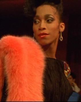 Octavia St. Laurent Is serious, has black hair with bangs, wearing silver earrings, an orange fur scarf, and a black dress with a big colored diamond on her left chest.