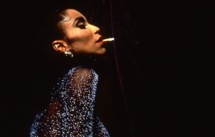 Octavia St. Laurent is serious, has black hair, and a cigarette in her mouth, wearing silver earrings and a black shimmering see-through dress.