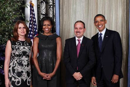 Standing in front of the USA Flag, Odeta Nishani, The First Lady of Albania's President (left) is smiling and has brown hair, her left hand at her back, wearing a black dress with a design. 2nd from left is Michelle Obama smiling, has black hair, both hands holding each other, and wearing a black shimmering dress. 3rd from left is Bujar Nishani smiling, has black hair and a mustache,  both hands holding each other, wearing white long sleeves, and a maroon necktie under a black suit. On right is Barack Obama smiling, has black hair, wearing white long sleeves, a blue necktie under a black suit.