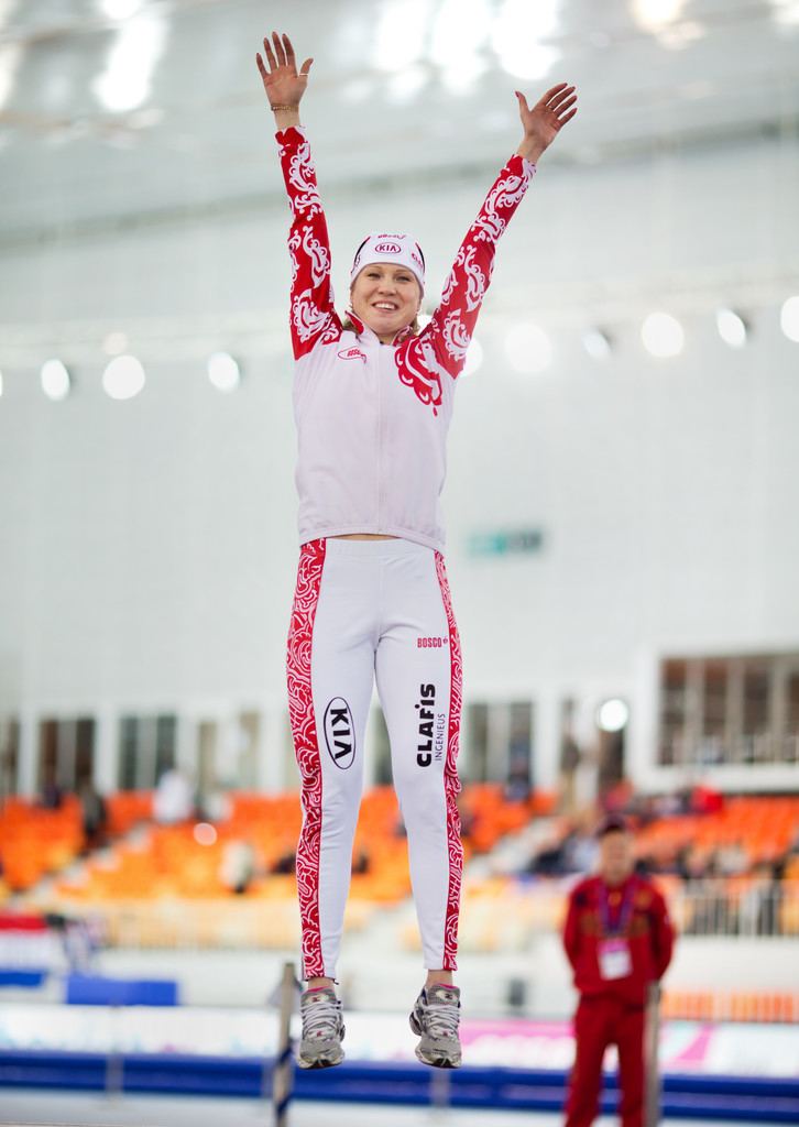 Olga Fatkulina smiling while jumping in the air with both of her hands raised inside the ice rink with people in the background, wearing a white headband with the printed logo "KIA", gray shoes, a red and white sleeved printed jacket, and leggings with the words "KIA, CLAFIS, and BOSCO" printed on it