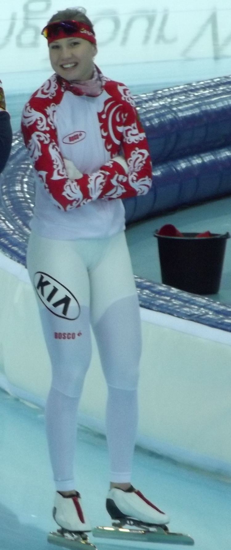 Olga Fatkulina smiling with folded arms and standing beside an ice rink and a black pail in the background, wearing white gloves, white leggings with prints, a red headband, sunglasses, and a red and white figure skates red and white long-sleeved jacket with prints