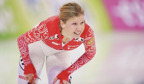 Olga Fatkulina smiling while bending forward with her hands on her knees and holding her sunglasses, having blonde hair, wearing earrings, a red printed jacket, and white leggings