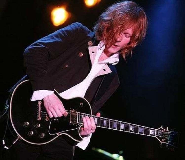 Oliver Thompson playing the guitar while wearing a black coat and white long sleeves