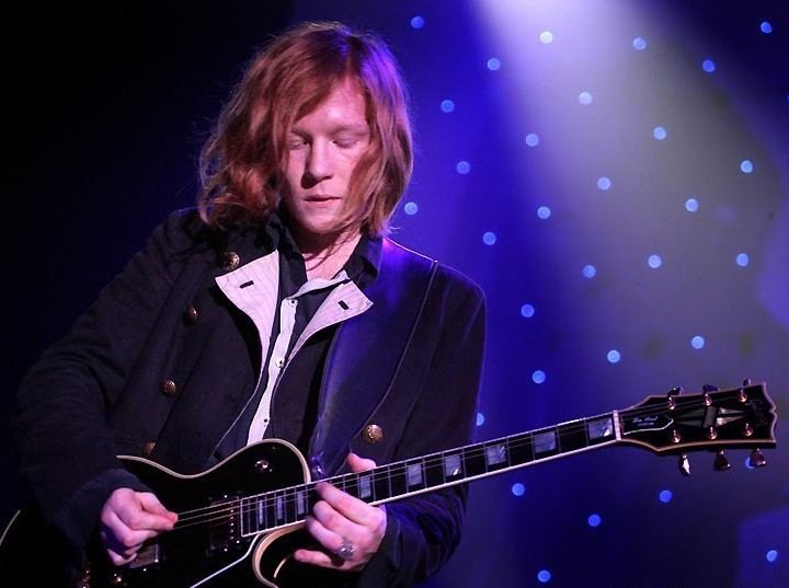 Oliver Thompson playing the guitar while wearing a black coat, black and white long sleeves, and ring