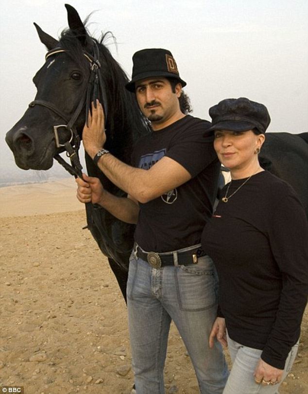 Omar bin Laden holding the horse' face while wearing a black hat, black printed shirt, and denim pants with his wife beside him