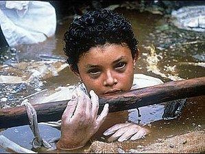 Omayra Sánchez with short curly hair, wearing a white shirt, holding a log, and soaked in the water.