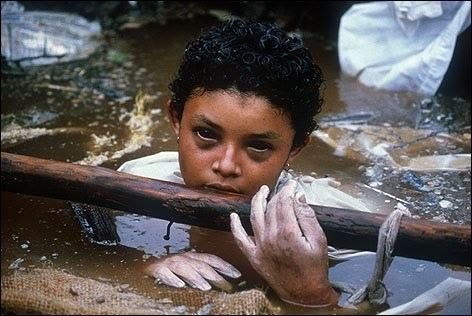 Omayra Sánchez with short curly hair, wearing a white shirt, holding a log, and soaked in the water.