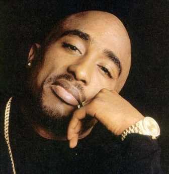 Tupac Shakur with a serious face while leaning on his hand, with beard and mustache, wearing earrings, a necklace, ring, watch, and a black long sleeve shirt.