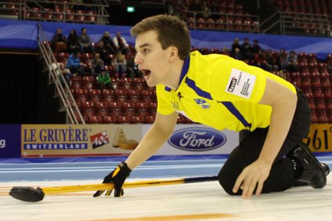 Oskar Eriksson kneeling on the floor with his mouth open while holding the curling broom and there are some people sitting on the bleachers. Oskar is wearing a yellow and blue t-shirt, black gloves, black pants, and black shoes