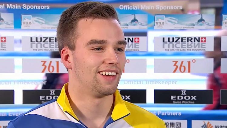 Oskar Eriksson smiling and looking at something, with different logos of brands in the background, while wearing a white, blue, and yellow jacket
