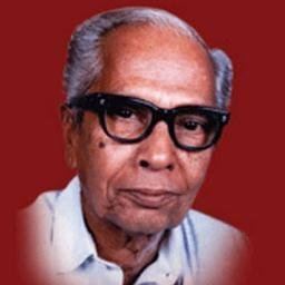 P N Panicker smiling with his gray hair on a red background, wearing black eyeglass and a white polo