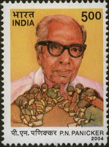In an Indian Postal stamp of Rs 5 is P N Panicker smiling with his gray hair in a photograph with women sitting and bowing their heads in a yellow background and a black wavy border, he is wearing black eyeglasses and a white polo.