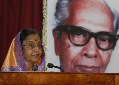 P N Panicker smiling with his gray hair and he is wearing black eyeglass in a photograph.. The Indian President Pratibha Patil speaking in a black microphone and has black hair with a bindi on her forehead wearing an eyeglass, and a purple and orange saree.
