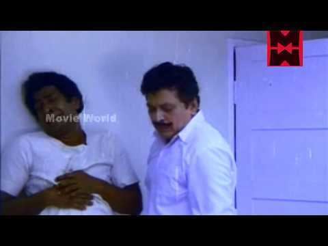 In the movie scene Paavam Krooran 1984, Captain Raju (left) is crying while holding his stomach with his hands together has black hair wearing a white top, Shankar Rs (left) standing while looking down has a mustache and black hair wearing a white long sleeve.