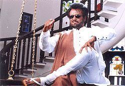 Rajinikanth sitting on the chair while wearing white long sleeves, white pants, black shades, and brown sash