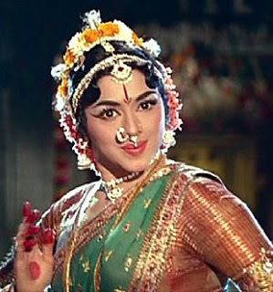 Padmini smiling while wearing a green and brown dress, nose jewel, headdress, and earrings