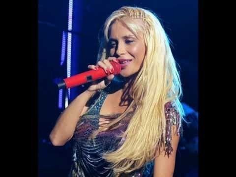 Paola Foka is smiling while singing and holding a red microphone, with long blonde hair and wearing a multi-colored sleeveless top.