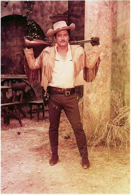 Paquito Diaz holding a rifle while wearing a cowboy hat, leather jacket, long sleeves, belt, pants, and brown shoes